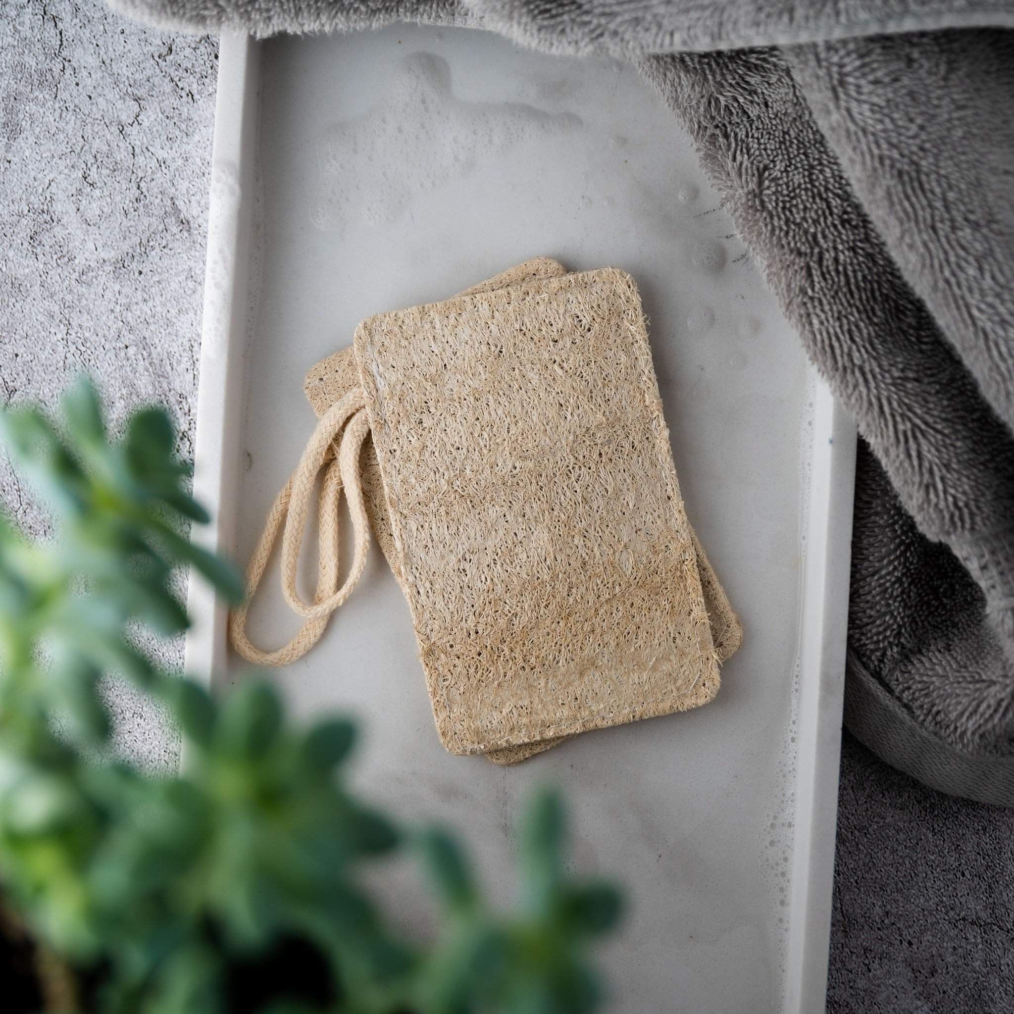 Natural Loofah Pad 2-Pack with 2x beige loofahs in frame with grey towels in side for aesthetic and white backrop