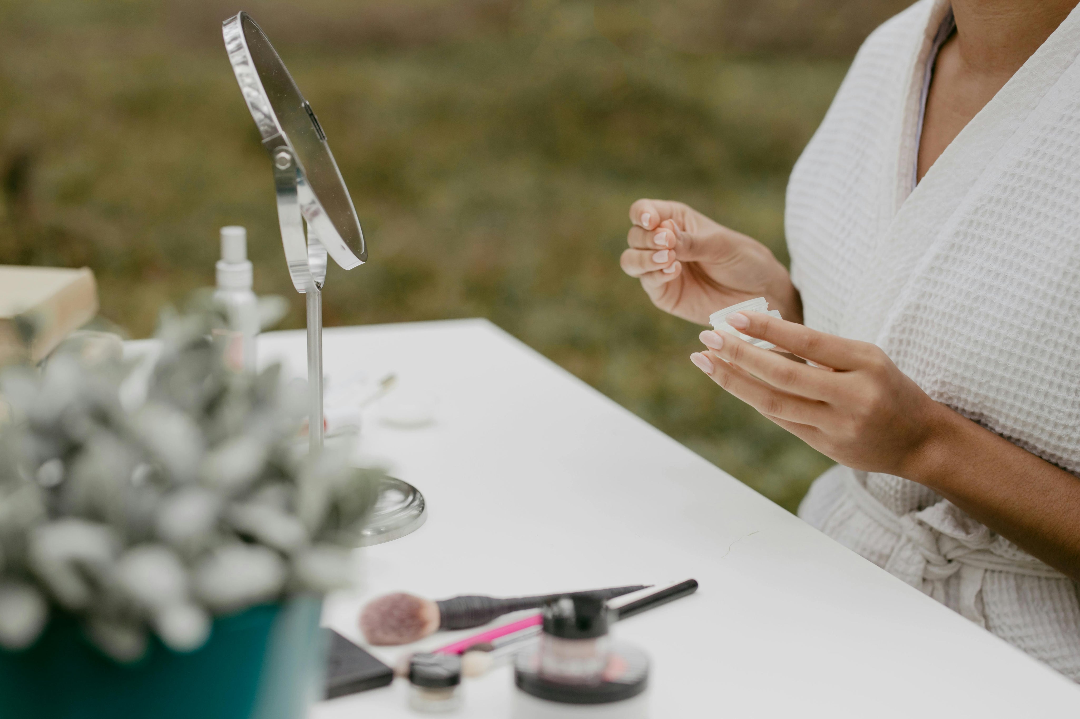 close up shot of female hands holding skincare product in a garden backdrop next to a table with mirror and makeup products